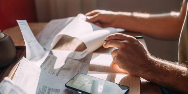 Close Up Of Man's Hands Reading Monthly Electric Bill And Using Phone As A Calculator