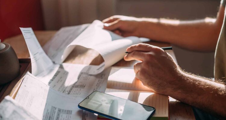 Close Up Of Man's Hands Reading Monthly Electric Bill And Using Phone As A Calculator